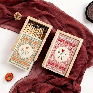 May include: Two wooden matchbox sets with decorative labels reading "The Perfect Match." One is open, revealing matches. The labels feature initials, a heart, and the date 08.25.2026. A small candle and decorative elements are also present.