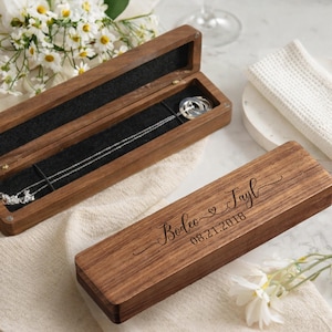 May include: Two wooden jewelry boxes, one open revealing a silver necklace. The closed box has engraved text: "Bodee & Tayl 08.21.2018". The boxes are on a white surface with a white cloth and flowers.