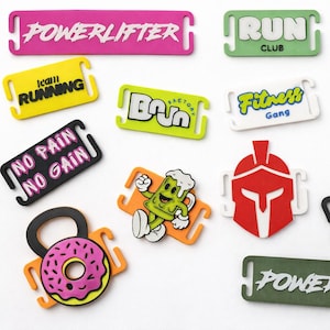 May include: A collection of colorful fitness-themed shoe charms. Designs include "POWERLIFTER", "RUN CLUB", "ATHLETE", "NO PAIN NO GAIN", and a weight with a donut. Various colors like pink, green, blue, yellow, and black.