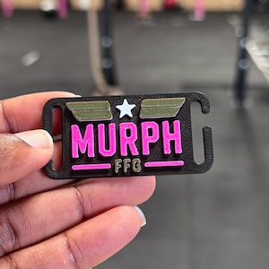 Peut inclure: Une étiquette rectangulaire en plastique noir avec des fentes à chaque extrémité, présentant le texte MURPH en lettres rose vif, les lettres FFG en texte rose plus petit, et deux barres vert olive entourant une étoile blanche au centre.