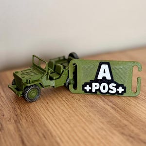 Peut inclure: Un jouet jeep vert olive à côté d'une étiquette verte assortie avec le texte "A +POS+" en blanc. La jeep a des roues détaillées et un design classique. L'étiquette a une forme incurvée avec un trou.