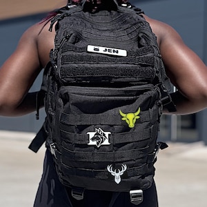 Peut inclure: Sac à dos tactique noir avec plusieurs compartiments et sangles MOLLE. Comprend une étiquette nominative blanche avec "JEN" et plusieurs écussons décoratifs, dont un loup, un taureau et un cerf.