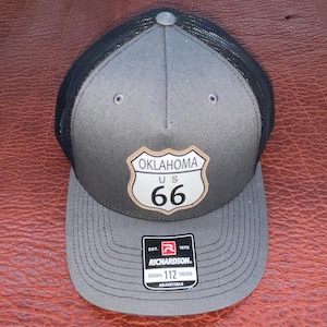 May include: A gray and black trucker hat with a curved brim. The hat features a patch with the Oklahoma US 66 route shield. The hat is adjustable and has a Richardson 112 tag.
