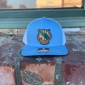 May include: A light blue trucker hat with a white mesh back. The front has a patch with a Native American design in brown, green, and red. The hat has a curved peak and a Richardson 112 tag.