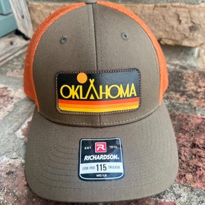 May include: A brown and orange trucker hat with the word "OKLAHOMA" in yellow and orange on a leather patch. The hat has a curved brim and a mesh back. The tag reads "Richardson 115 Trucker".
