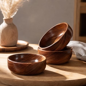 Peut inclure: Trois bols ronds en bois avec une finition brun foncé et des motifs de grain de bois visibles. Un bol est placé au premier plan, tandis que deux autres bols sont empilés l'un sur l'autre derrière, mettant en valeur la texture lisse du bois.