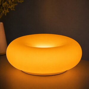 May include: A circular, donut-shaped table lamp emitting a warm, soft yellow glow. The lamp features a smooth, matte surface that diffuses light evenly across its rounded, ring-like structure, creating a bright, illuminated center aperture.