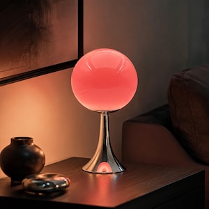 Peut inclure: Une lampe de table composée d'un abat-jour sphérique en verre rouge lumineux, reposant sur une base en métal argenté poli et réfléchissant, dotée d'un pied évasé en forme de trompette sur un socle circulaire plat.