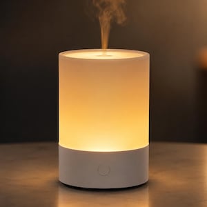 Può includere: Un diffusore di oli essenziali cilindrico bianco con una parte superiore in vetro smerigliato che emette una luce calda e un sottile getto di vapore. La base è di colore bianco solido e presenta un pulsante di accensione circolare sul davanti.