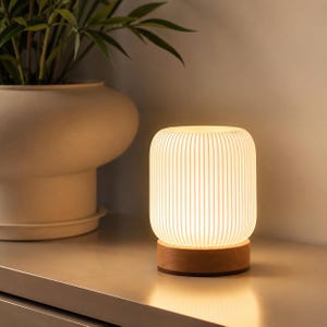 Peut inclure: Une lampe de table cylindrique dotée d'un abat-jour blanc à côtes verticales qui émet une lueur chaude et douce. La lampe repose sur une base circulaire en bois aux tons naturels, offrant une assise stable à ce luminaire.