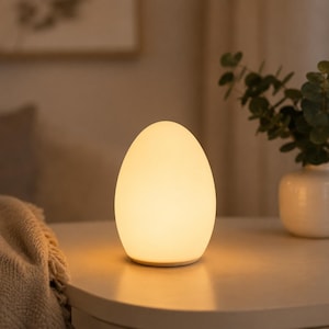 May include: A smooth, egg-shaped table lamp emitting a soft, warm, diffused glow. The lamp has a solid, opaque white exterior surface and a rounded, symmetrical silhouette that rests flat on a surface, providing ambient illumination.