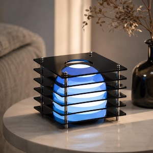 Peut inclure: Une lampe carrée composée d'une pile de cinq plaques en acrylique noir maintenues par quatre tiges métalliques argentées. Une source lumineuse sphérique bleue et blanche brille au centre, créant un effet superposé à travers le cadre.