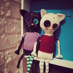 May include: Two knitted cat toys with mismatched eyes and striped legs. The black cat is wearing a purple shirt and the white cat is wearing a red shirt.
