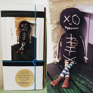 May include: A knitting kit to make a skeleton doll. The kit includes yarn, needles, stuffing, and a pattern. The doll is black with white stitching and has a heart on its chest. The doll is sitting on a wooden surface.