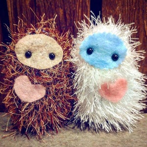 Op de afbeelding: Twee pluche, handgemaakte knuffels met harten. Eén is bruin met een beige gezicht en de andere is wit met een blauw gezicht.