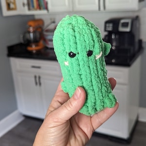 Könnte beinhalten: Ein grünes, gestricktes Kaktusspielzeug mit gestickten Augen und einem weißen, gestickten Lächeln. Der Kaktus wird in einer Hand gehalten.
