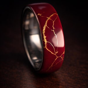 Puede incluir: Un anillo ancho y rojo con un patrón de craquelado dorado. El anillo tiene un acabado pulido y un interior plateado. Las vetas doradas crean un contraste llamativo con el rojo. Una joya única.