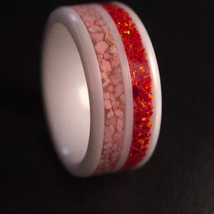 May include: A white ring with a central band of pink stones and a band of red, sparkling stones. The ring has a smooth, polished white exterior. The gemstones are set within the ring's design, creating a striking contrast.