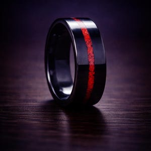 May include: A black ring with a vibrant red inlay. The ring has a polished, reflective surface, and the red inlay is a contrasting stripe. The ring is displayed on a dark wooden surface, creating a striking visual.