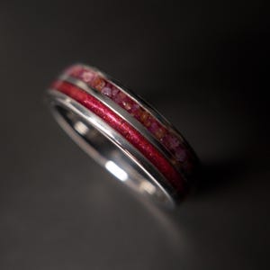 May include: A silver ring with a vibrant red enamel center band and a row of small, reddish-pink gemstones on either side. The ring's polished metal reflects light, creating a striking contrast against the dark background. A close-up shot highlights the ring's intricate design.