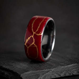 Può includere: Un anello caratterizzato da una fascia esterna rosso intenso con un evidente motivo a crepe color oro che attraversa la superficie. L'interno dell'anello presenta una finitura metallica scura e liscia, in contrasto con l'esterno rosso e oro.