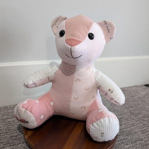 May include: A stuffed bear toy constructed from various fabric patches in shades of pink and white. The bear features black button eyes, a stitched nose and mouth, and patterns including polka dots, stripes, rainbows, and small flowers.