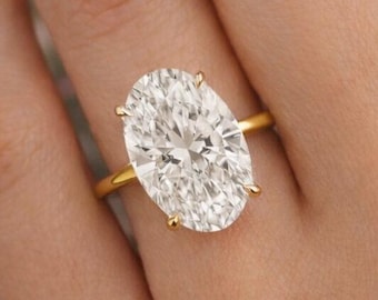 May include: A gold ring with a large, oval-cut diamond. The diamond is set in a four-prong setting. The ring is on a person's finger, with a floral background.