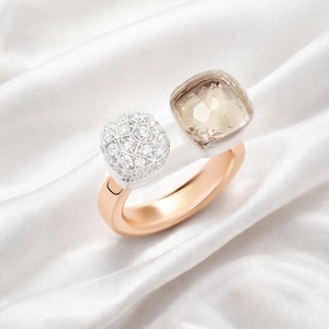 Puede incluir: Un anillo abierto con una banda de oro rosa y dos engastes distintos. Un lado sostiene una piedra preciosa cuadrada de color marrón claro facetada, mientras que el otro lado presenta un grupo cuadrado de pequeñas piedras claras talladas en plata.