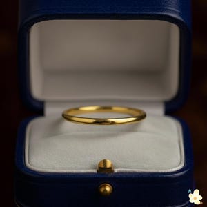 May include: A gold wedding band sits inside a blue jewelry box with a white interior. The ring is simple and elegant, with a smooth, polished surface. The box is closed with a gold-colored clasp.