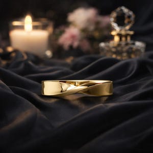 May include: A gold bracelet with a twisted design. The bracelet is made of a shiny metal and is displayed on a black, silky fabric. A lit candle and a decorative perfume bottle are in the background.