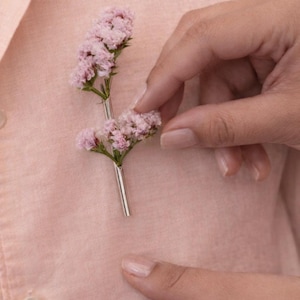 以下が含まれることがあります： 小さなピンクの花が銀色の茎に付いた、繊細な花のブローチ。 ブローチは薄いピンク色のシャツにピンで留められており、柔らかくエレガントな美しさを演出しています。 デザインはシンプルで洗練されており、自然にインスパイアされた魅力を添えるのに適しています。