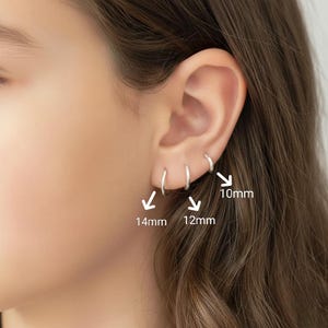 Puede incluir: Primer plano de una oreja con tres pendientes de aro plateados de 10 mm, 12 mm y 14 mm de diámetro. Los pendientes son sencillos y brillantes. La imagen está bien iluminada, mostrando los detalles de las joyas.