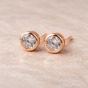 May include: A pair of rose gold stud earrings. Each earring features a round, clear, faceted gemstone set in a bezel setting. The earrings are displayed on a light, textured surface.