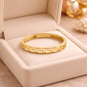 May include: A gold-colored ring with a Greek key design, resting in a beige jewelry box. The ring's intricate pattern is visible, and the overall aesthetic is elegant and classic. The ring is a piece of jewelry.