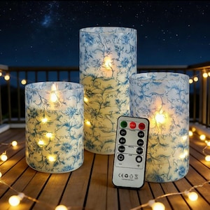 May include: Set of three battery-operated LED pillar candles with a blue floral design. The candles are different heights and include a remote control. The candles are on a wooden surface with string lights.