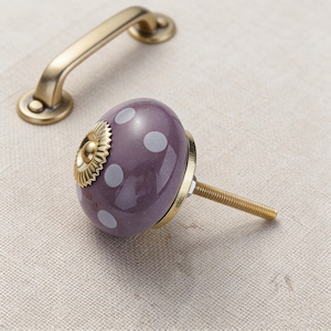 May include: A decorative cabinet knob with a purple, polka-dotted ceramic design and a gold-coloured metal base. A matching gold-coloured handle is in the background. The knob has a threaded rod for installation.