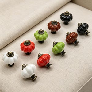 May include: A collection of ceramic cabinet knobs in various colors, including white, red, green, brown, and black. Each knob has a pumpkin-like shape with a bronze-colored base and top. The knobs are arranged on a neutral-toned surface.