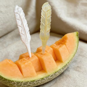 Puede incluir: Un melón cantalupo cortado por la mitad con pulpa naranja en cubos, servido con dos palillos decorativos en forma de pluma. Uno es blanco, el otro es amarillo pálido. El melón está sobre una superficie de tono neutro.