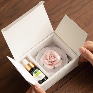 May include: A white gift box with a rose-shaped decorative item and a small bottle of essential oil. The rose is pink and encased in a clear dome. The oil bottle has a gold cap and a label with text.