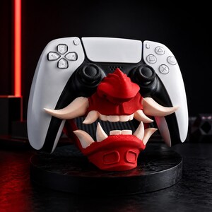 May include: A white and black video game controller featuring a red and cream-colored oni mask attachment. The mask covers the center of the controller, displaying prominent fangs, a nose, and a mouth, integrated into the controller's design.