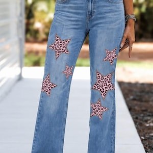 May include: Light blue denim jeans featuring a high-waisted fit and a straight-leg silhouette. The jeans are decorated with multiple five-pointed star patches in a pink and black leopard print pattern distributed across the front of the legs.