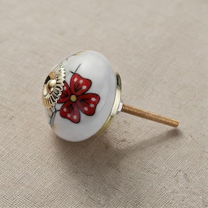 May include: White ceramic cabinet knob with a red flower design. The knob features a gold-colored metal base and a threaded rod for installation. The flower has red petals with white dots and a yellow center.