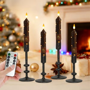 Puede incluir: Juego de cuatro velas LED a pilas con mando a distancia. Las velas son de color marrón oscuro con pequeñas luces y están colocadas en soportes de metal negro. El mando a distancia es blanco. Un árbol de Navidad y una chimenea en el fondo.
