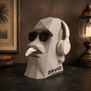 May include: A light grey, geometric, faceted head-shaped tissue dispenser featuring the name DAVID printed on the side. The sculpture wears dark-rimmed sunglasses and white over-ear headphones, with a white tissue protruding from the mouth area.
