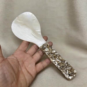 Puede incluir: Una cuchara de arroz decorativa con una pala nacarada blanca y un mango con incrustaciones de pequeños trozos de forma irregular en tonos marrón, beige y blanco. La cuchara está diseñada para servir arroz. La longitud de la cuchara es de unos 20 cm.