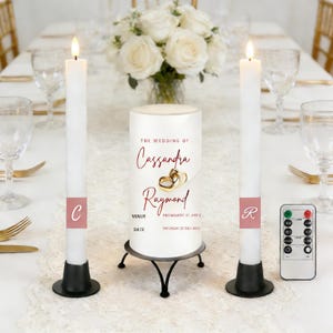 May include: A wedding table setting with two white pillar candles, each with a black base, flanking a central candle. The central candle displays wedding details. A remote control is also visible.