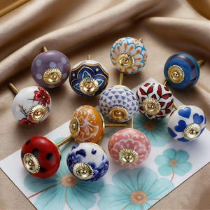 May include: A collection of decorative ceramic cabinet knobs in various colors and patterns. The knobs feature designs such as polka dots, floral motifs, and geometric shapes. Each knob has a gold-colored base and stem, adding a touch of elegance.