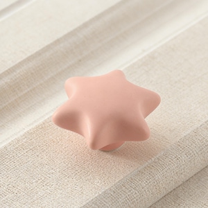 May include: A light pink, star-shaped cabinet knob. The knob is made of a smooth material and has a rounded, ergonomic design. The knob is shown on a light-colored, textured surface, suggesting a home decor or furniture item.