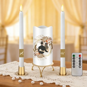 May include: A wedding candle set featuring a central pillar candle with a couple illustration and two tall, battery-operated candles. The candles are white with gold accents and a remote control. The set is decorated with floral elements.