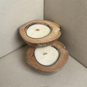 May include: Two coconut shell candles with creamy white wax and wooden wicks. The candles are nestled on a neutral-toned surface, creating a natural and relaxing aesthetic. These candles are a unique home decor item.